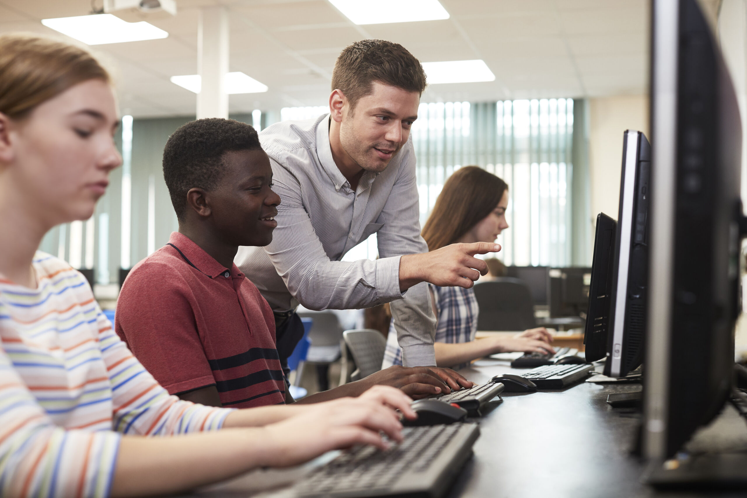 Teacher Helping Male High School Student Working In Computer Class